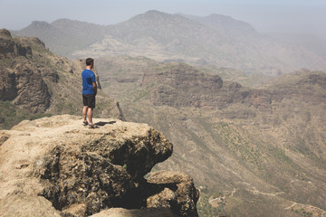 Fototapeta premium Tourist on mountain edge facing great landscape views. Young man on blue t shirt standing on top of rock on sunny day in Roque Nublo. Adventure, explore, freedom, visionary, solitude concepts
