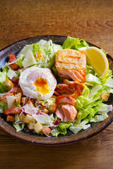 Salmon caesar salad. Crispy pan fried salmon fillet, bacon, poached egg, romaine lettuce and croutons in bowl on wooden table. vertical