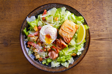 Salmon caesar salad. Crispy pan fried salmon fillet, bacon, poached egg, romaine lettuce and croutons in bowl on wooden table. overhead, horizontal