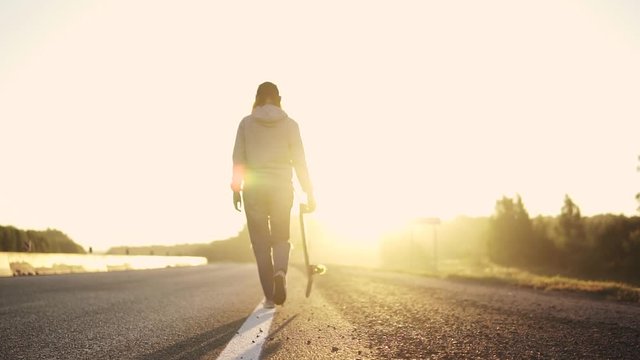 silhouette of a girl with a skateboard in the hands of a road running along the roadside at sunset. back view. slow motion.