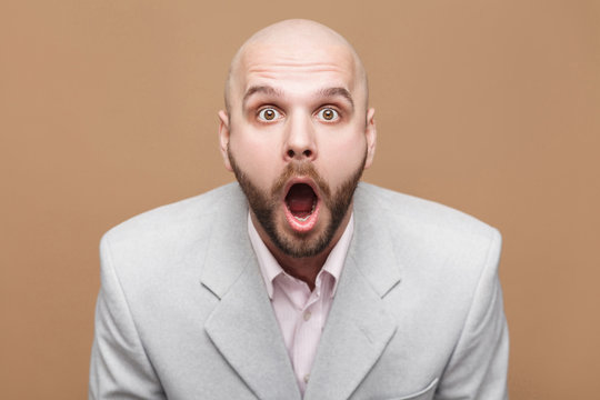 Pointing finger and amazed portrait of handsome middle aged businessman in classic light gray suit standing, looking at camera, wondering and showing brown background. indoor studio shot, isolated.