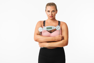 Young woman with excess weight in sporty top holding scales in hands while thoughtfully looking in camera over white background. Plus size model