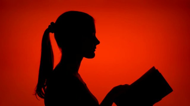 Silhouette of young woman reading physical print book on red background. Female's face in profile studying. Black contur shadow of teenager's half-face. Concept of old traditional classical paper