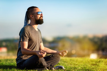 Portrait of bearded meditating man sitting at green grass in lotus pose at city sunset background.