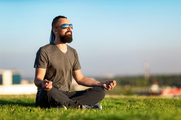 Portrait of bearded meditating man sitting at green grass in lotus pose at city sunset background.