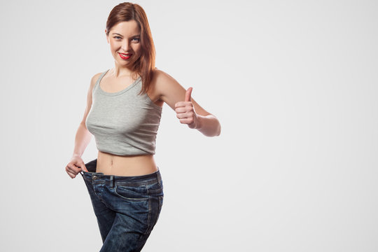 portrait of happy beautiful slim waist of young woman standing in big jeans and gray top showing successful weight loss, indoor, studio shot, isolated on light gray background, diet concept.