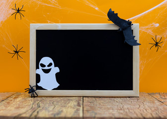 Halloween background concept. Blank blackboard with decor paper cut funny ghost faces, cobweb on wooden table and orange backdrop