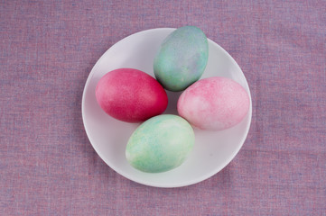 four multicolored painted Easter eggs on a white plate and lilac tablecloth. holiday easter greeting card, poster, wallpaper