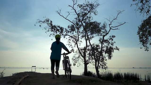 Silhouette of cyclist woman walking with bicycle at seaside, stretching exercise.