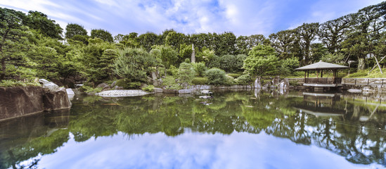 Fototapeta na wymiar Hexagonal Gazebo in the central pond of Mejiro Garden where ducks are resting and which is surrounded by large rocks and stone pagoda under the foliage of the pines trees and Momiji maple trees.