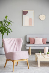 Pink armchair next to table in grey apartment interior with poster above couch. Real photo