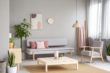 Wooden table on carpet and armchair in grey flat interior with plant next to sofa. Real photo