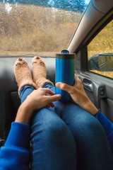 Feet in warm cute socks on car dashboard. Travel, road trip and autumn fall concept. Focus on thermos bottle cup with hot drink in female hands.