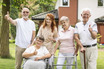 Thumbs up shown by happy female and male seniors and a caregiver outside their nursing home.