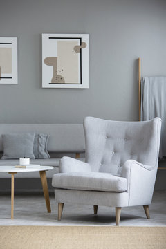 Grey armchair next to table in living room interior with posters above sofa with pillows. Real photo