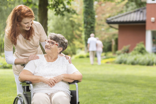 A Female Volunteer Helping A Disabled Elderly Woman In A Wheelchair In The Garden Outside A Private Luxurious Retirement Home. Blurred Background.