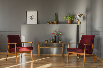 Flowers on wooden table between dark red armchairs in grey living room interior with poster. Real photo