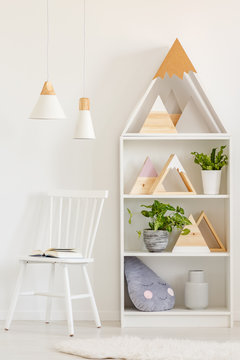 Open book on a simple, wooden chair and a bookcase with plants and DIY mountains decorations in a white, scandinavian living room interior