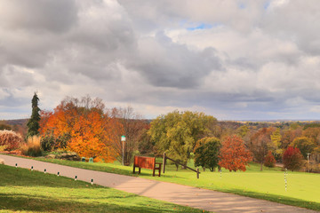 Beautiful fall nature background. Spectacular autumn rural cloudscape with colorful trees on a golf course. Path and wooden bench on a foreground. Leisure activity and lifestyle background.