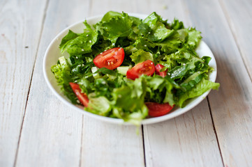 Salad from tomato, cucumbers, lettuce leaves and seeds of sesame on a white plate on a wooden background