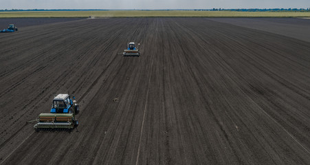 Aerial view of two blue tractors plows the earth in field on a summer day against a black earth...