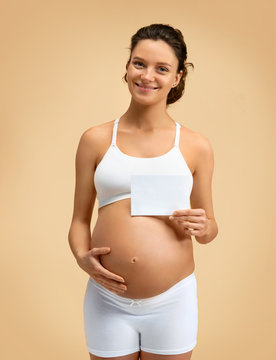 Happy pregnant woman touching her belly and holding greeting card on beige background. Pregnancy, maternity, preparation and expectation concept