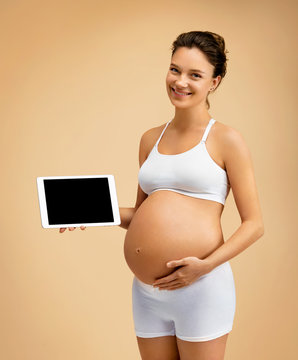 Happy pregnant woman holding tablet and touching her belly on beige background. Pregnancy, maternity, preparation and expectation concept