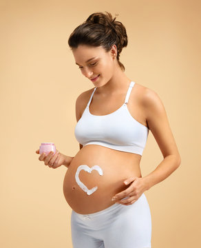 Smiling pregnant woman applying moisturizing cream on her belly on beige background. Pregnancy, maternity, preparation and expectation concept