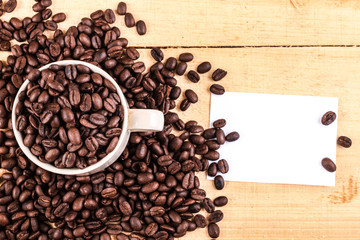 Cup of coffee full of coffee beans with blank sticky note on wooden background, top view