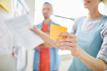 Close-up of positive manager holding adhesive note while planning project development with colleague and presenting her idea at meeting