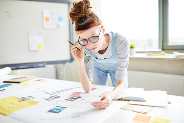 Serious thoughtful female hipster marketer adjusting glasses while reading notes on paper, she...