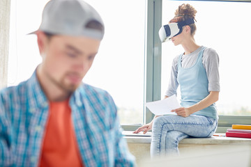 Fototapeta premium Pensive young female student in VR headset sitting on window-sill and using laptop while examining data, she working on project