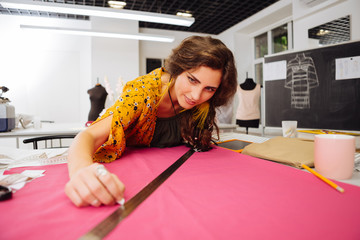 Attention. Smart cheerful dressmaker smiling and leaning on the table while holding a piece of white chalk and drawing the line on pink fabric