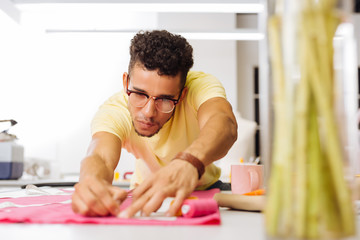 Careful man. Careful attentive young tailor wearing glasses and leaning to the table while holding piece of chalk and drawing lines on textile