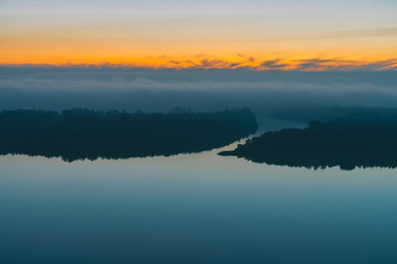 Fototapeta na wymiar Early blue sky reflected in river water. Riverbank with forest under predawn sky. Yellow stripe in picturesque sky. Fog hid trees on island. Mystical morning atmospheric landscape of majestic nature.