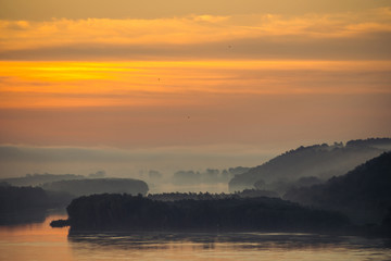 Morning haze above valley of river. Gold glow from dawn in sky and reflex on water. Birds flying in sky at sunrise. Fog on riverbank with forest. Colorful atmospheric landscape of majestic nature.