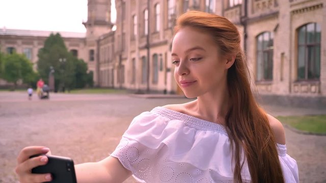 Beautiful ginger young woman with bare shoulders taking selfie with her phone and smiling, standing on street with building in background