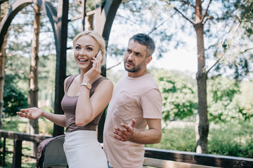 Fototapeta premium mother talking by smartphone near baby carriage in park, angry husband looking at her