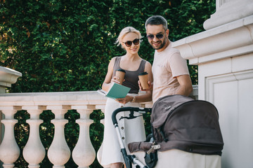 parents standing near baby carriage in park and holding book with coffee to go