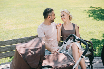 Obraz premium smiling parents sitting on bench near baby carriage in park and looking at each other