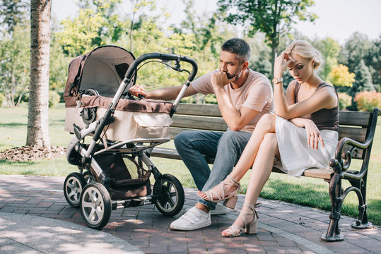 sad parents sitting on bench near baby carriage in park