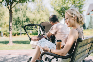 Fototapeta na wymiar side view of wife sitting near baby carriage with coffee to go and travel newspaper