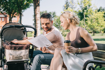 Naklejka premium wife showing newspaper to husband on bench near baby carriage in park