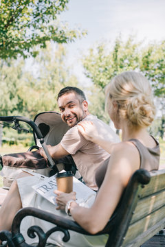 wife sitting near baby carriage with coffee to go and travel newspaper and looking at husband
