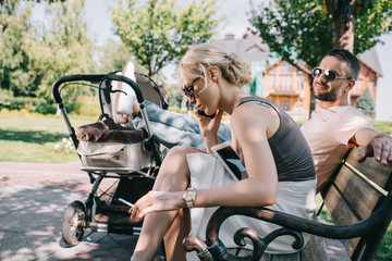 mother smoking cigarette near baby carriage in park and talking by smartphone