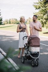 smiling parents walking with baby carriage and ice cream in park