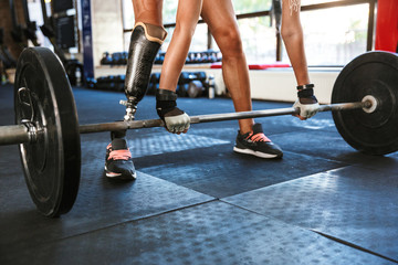 Fototapeta na wymiar Cropped image of athletic disabled sportswoman wearing prosthesis in tracksuit, training and lifting barbell in gym