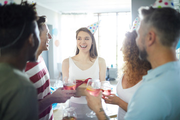 Obraz premium Cheerful laughing girl in birthday cap holding giftbox with present from her friends while they toasting with drinks