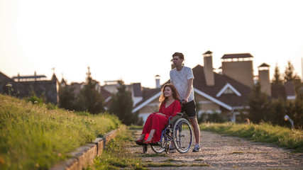 A beautiful girl in a wheelchair and her young man are walking in the park, next to beautiful houses