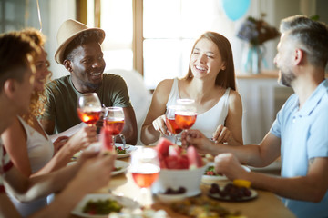 Joyful multi-ethnic friends sitting by festive table, having drinks and talking at home party